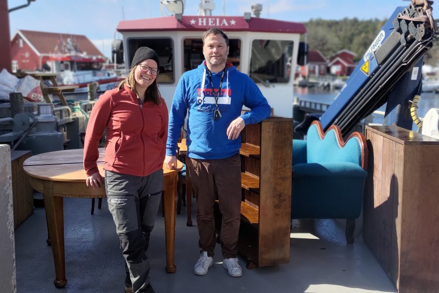 Bilde av to personer som står på en båt med en blå sofa og andre møbler