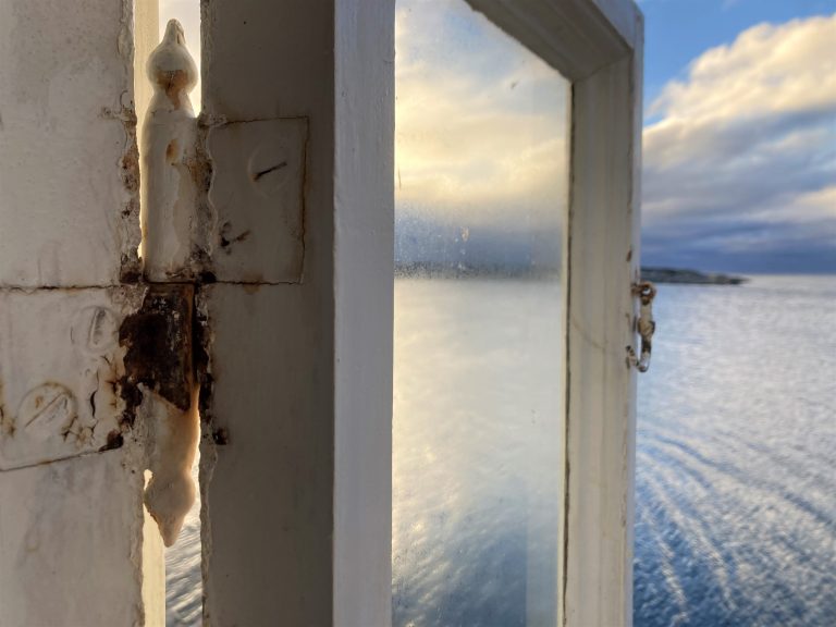 Et nærbilde av en vindushengsel som er gammel og rusten. I bakgrunnen ser man havet.