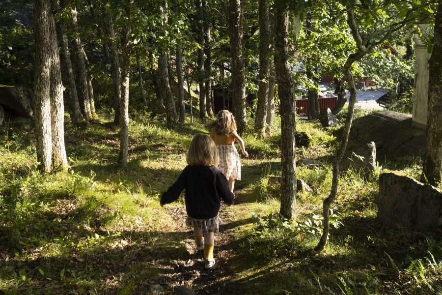 To små jenter løper på en sti i en skog. Trærne lager skygger over stien, som ellers ligger i sollys.