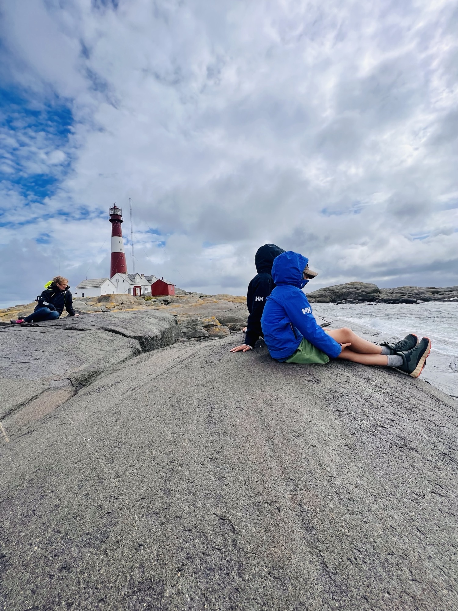 Tre personer sitter på et svaberg og ser utover mot havet. I bakgrunnen er et fyrtårn.