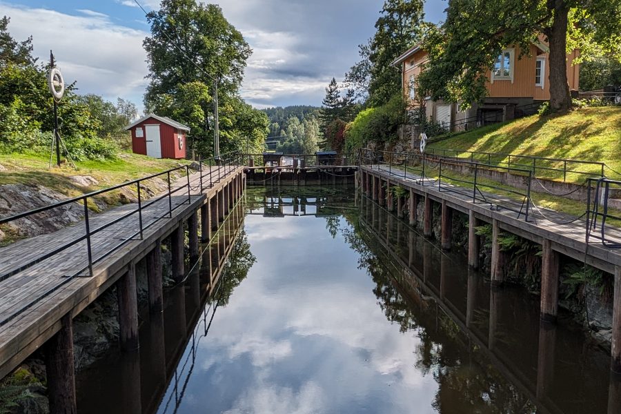 Bilde av et slusekammer fylt med vann. På den ene siden av slusa ligger en gul bygning. På den andre siden av slusa ligger en liten rød bod. Himmelen speiler seg i vannet i slusekammeret.