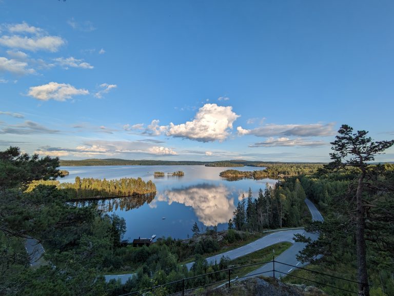 Bilde av et vann, sett fra en ås. En vei går i svinger langs vannet. Skyer speiler seg i vannet.