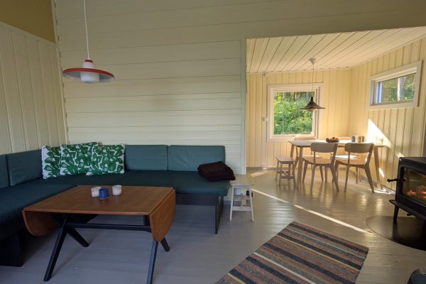 Bilde av en lys, gul stue med en grønn sofa i det ene hjørnet. Til høyre en vedovn med fyr i, og i bakgrunnen et spisebord og stoler i lyst treverk.
