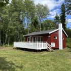 Bilde av en liten, rød hytte med en hvit terrasse foran. I forkant er det en grønn gressplen, og i bakgrunnen er det skog.