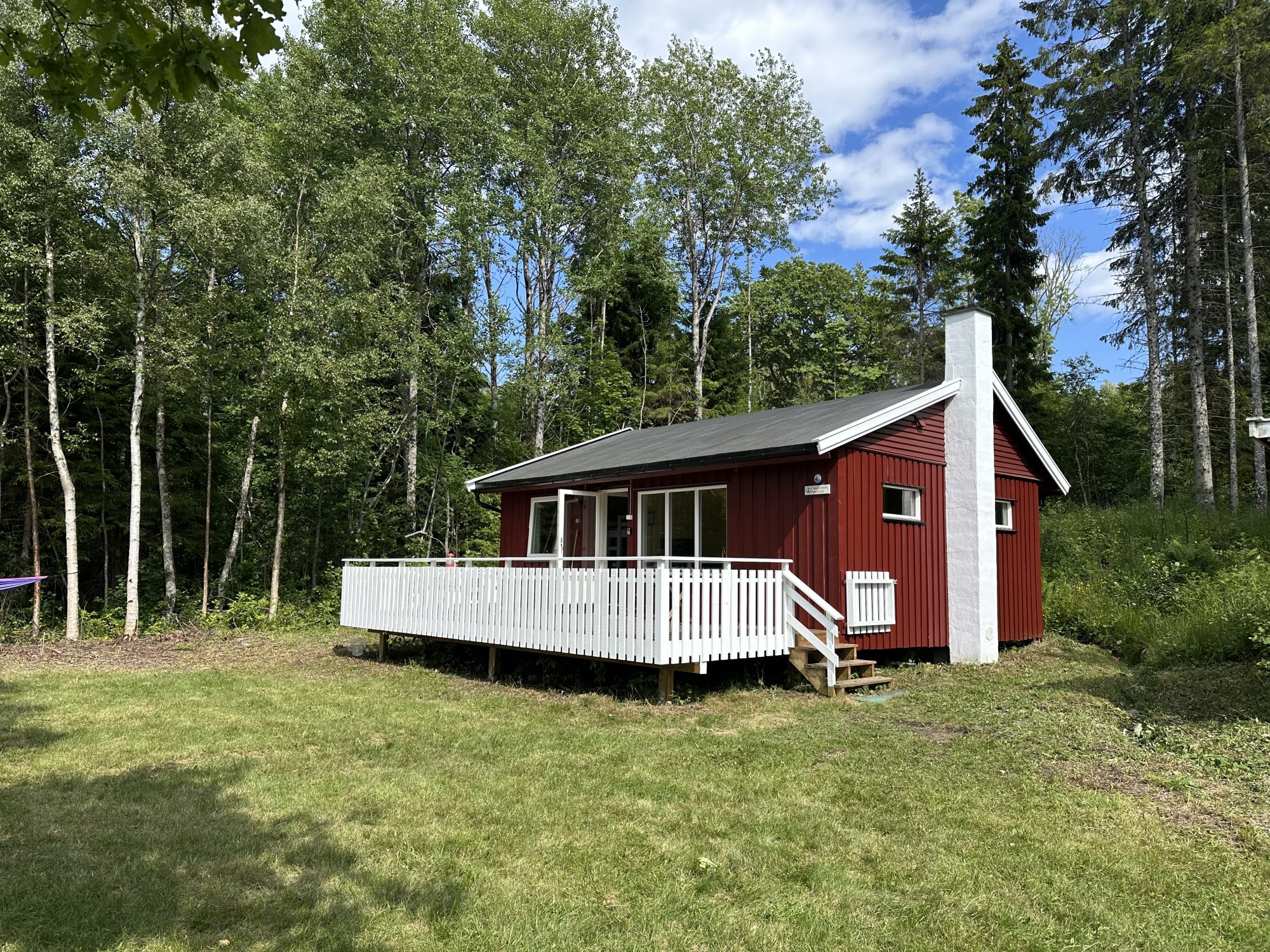 Bilde av en liten, rød hytte med en hvit terrasse foran. I forkant er det en grønn gressplen, og i bakgrunnen er det skog.