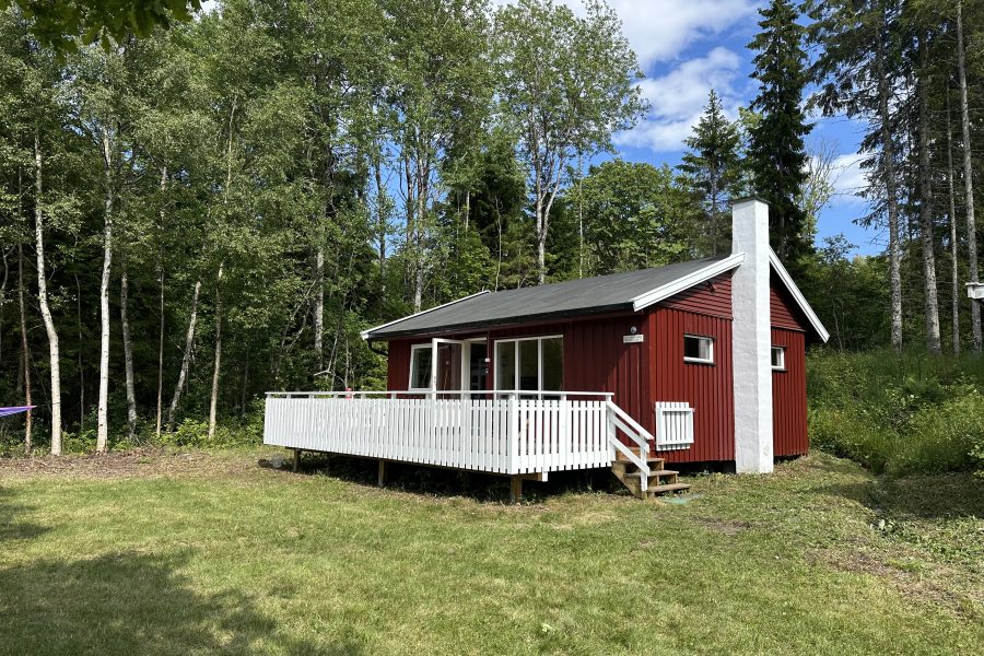 Bilde av en liten, rød hytte med en hvit terrasse foran. I forkant er det en grønn gressplen, og i bakgrunnen er det skog.
