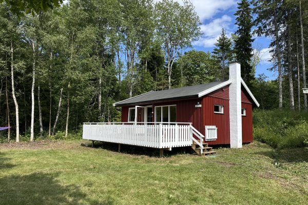 Bilde av en liten, rød hytte med en hvit terrasse foran. I forkant er det en grønn gressplen, og i bakgrunnen er det skog.