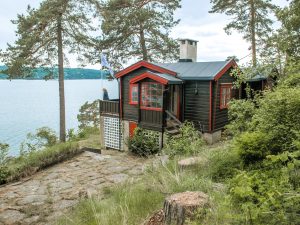 På bildet er det en brun, laftet hytte. Nedenfor hytta skråner terrenget ned mot sjøen, som man ser i bakgrunnen. På verandaen til hytta står en dame og ser ut over sjøen.