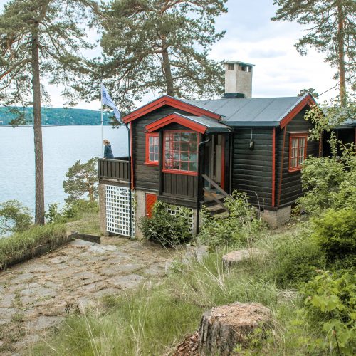 På bildet er det en brun, laftet hytte. Nedenfor hytta skråner terrenget ned mot sjøen, som man ser i bakgrunnen. På verandaen til hytta står en dame og ser ut over sjøen.