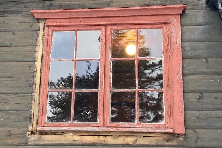 Et bilde av et vindu som har ødelagte karmer. Den røde malinga som er igjen på karmene flasser og er falmet, og det er tydelig at vinduet trenger vedlikehold.