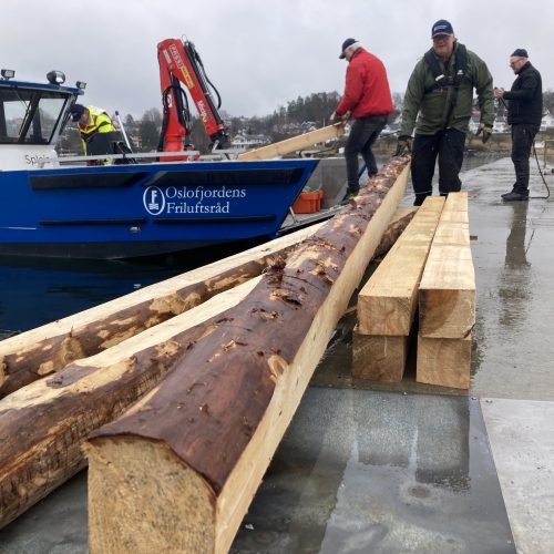 På dette bildet ser man store tømmerstokker som ligger på en brygge. Inntil brygga er det en båt, og fire menn er opptatt med å få flere stokker av båten.