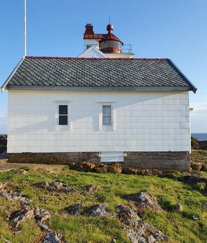 Bilde av en hvit bygning med grått skifertak. Foran bygningen er det grønt gress, og bak bygningen er himmelen blå. Man kan også se en fyrlykt i bakgrunnen. Fra en flaggstang vaier en vimpel i vinden.