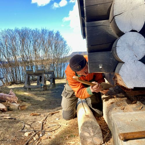 På bildet ser man hjørnet på en laftet hytte. Man kan se at en av stokkene mangler, og en mann sitter på knærne med en øks i hånda.