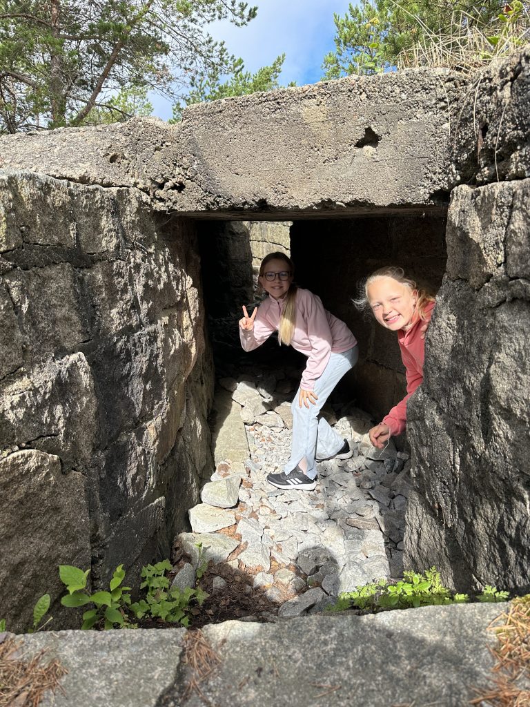 To jenter står med bøyd rygg i en gammel bunkers. De smiler mot kamera.