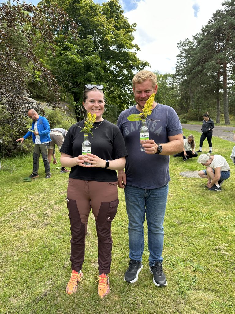 En mann og en dame står i gresset og viser fram hver sin bukett. I bakgrunnen er det flere mennesker som plukker opp ting fra bakken.