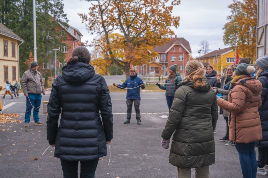 En gruppe mennesker står i ring på en plass mellom flere bygninger. I hendene sine holder de hvert sitt meterlange tau. I bakgrunnen er det noen lekeapparater.