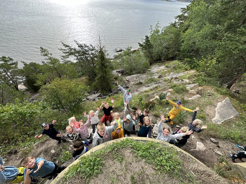 Et bilde av mange barn som står ute ved et vann og strekker hendene i været.