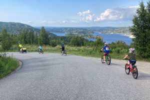 En voksen og fire barn sykler nedover en bakke. I bakgrunnen ser man fjorden.